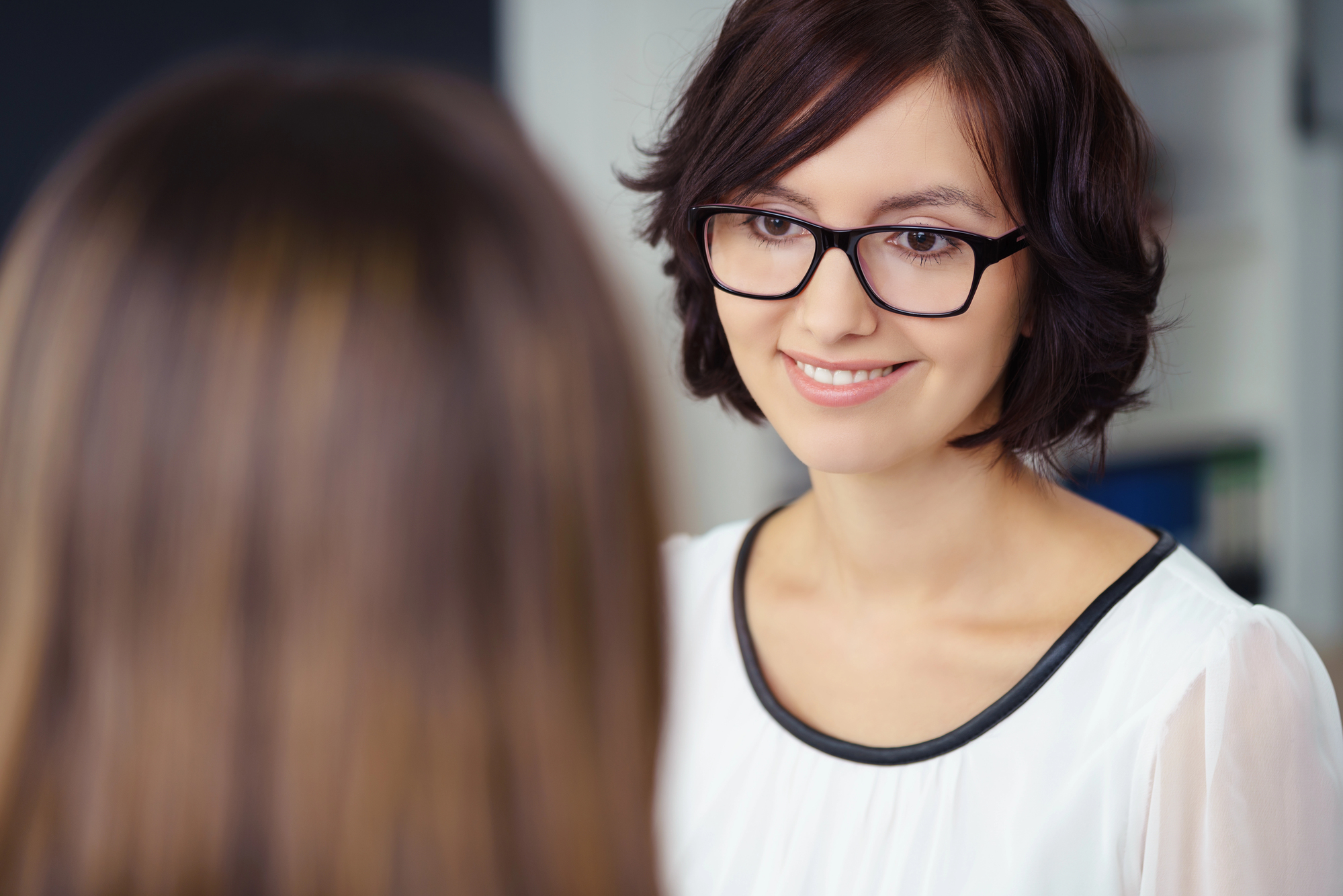 A woman with shoulder-length brown hair and glasses is smiling while talking to another person who is facing away from the camera. She is wearing a white blouse with black trim, and the background appears to be a blurred indoor setting.