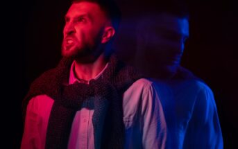 A man with a beard, wearing a scarf and button-up shirt, stands against a dark background. His expression appears intense with a red and blue light effect, creating a blurred double-exposure of his profile.