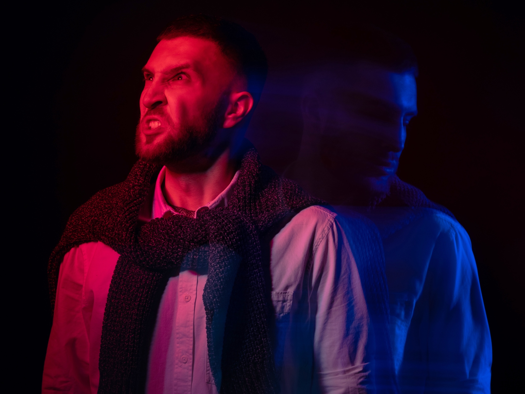 A man with a beard, wearing a scarf and button-up shirt, stands against a dark background. His expression appears intense with a red and blue light effect, creating a blurred double-exposure of his profile.