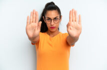 A woman with glasses and a ponytail stands against a white background, wearing an orange shirt. She extends both hands forward with open palms, signaling to stop. Her expression is serious and assertive.