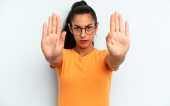 A woman with glasses and a ponytail stands against a white background, wearing an orange shirt. She extends both hands forward with open palms, signaling to stop. Her expression is serious and assertive.