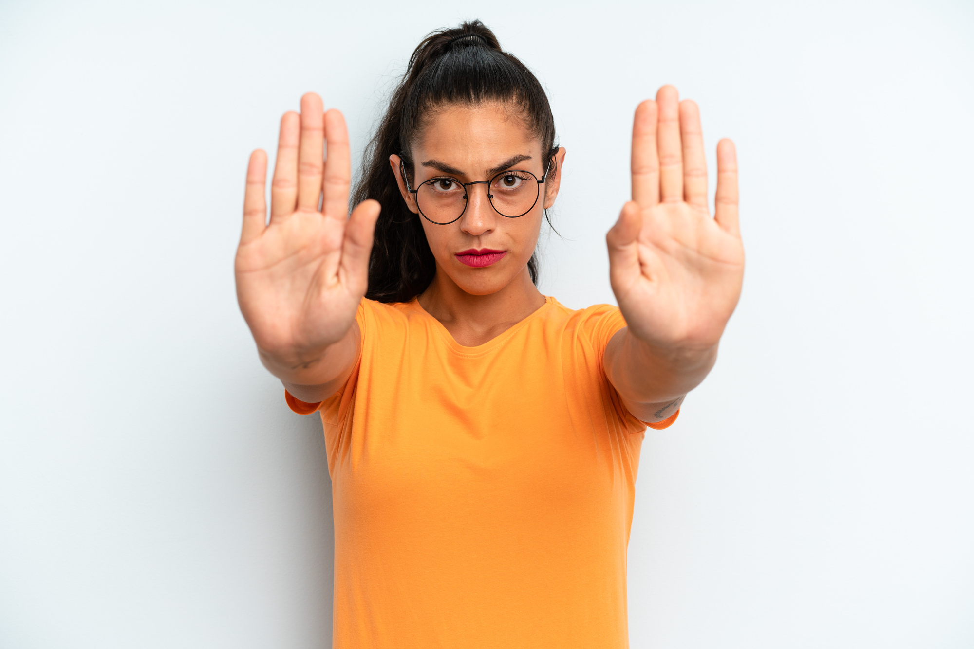 A woman with glasses and a ponytail stands against a white background, wearing an orange shirt. She extends both hands forward with open palms, signaling to stop. Her expression is serious and assertive.