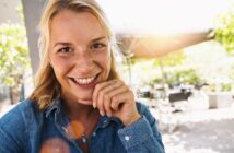 A woman with blonde hair smiles warmly at the camera. She's wearing a blue denim shirt and sitting outdoors, with sunlight filtering through green foliage in the background. She rests her chin on her hand, creating a relaxed, cheerful atmosphere.