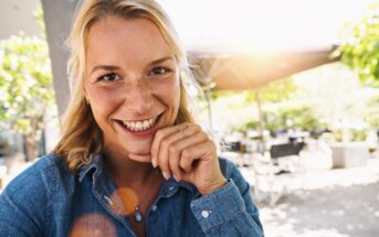 A woman with blonde hair smiles warmly at the camera. She's wearing a blue denim shirt and sitting outdoors, with sunlight filtering through green foliage in the background. She rests her chin on her hand, creating a relaxed, cheerful atmosphere.