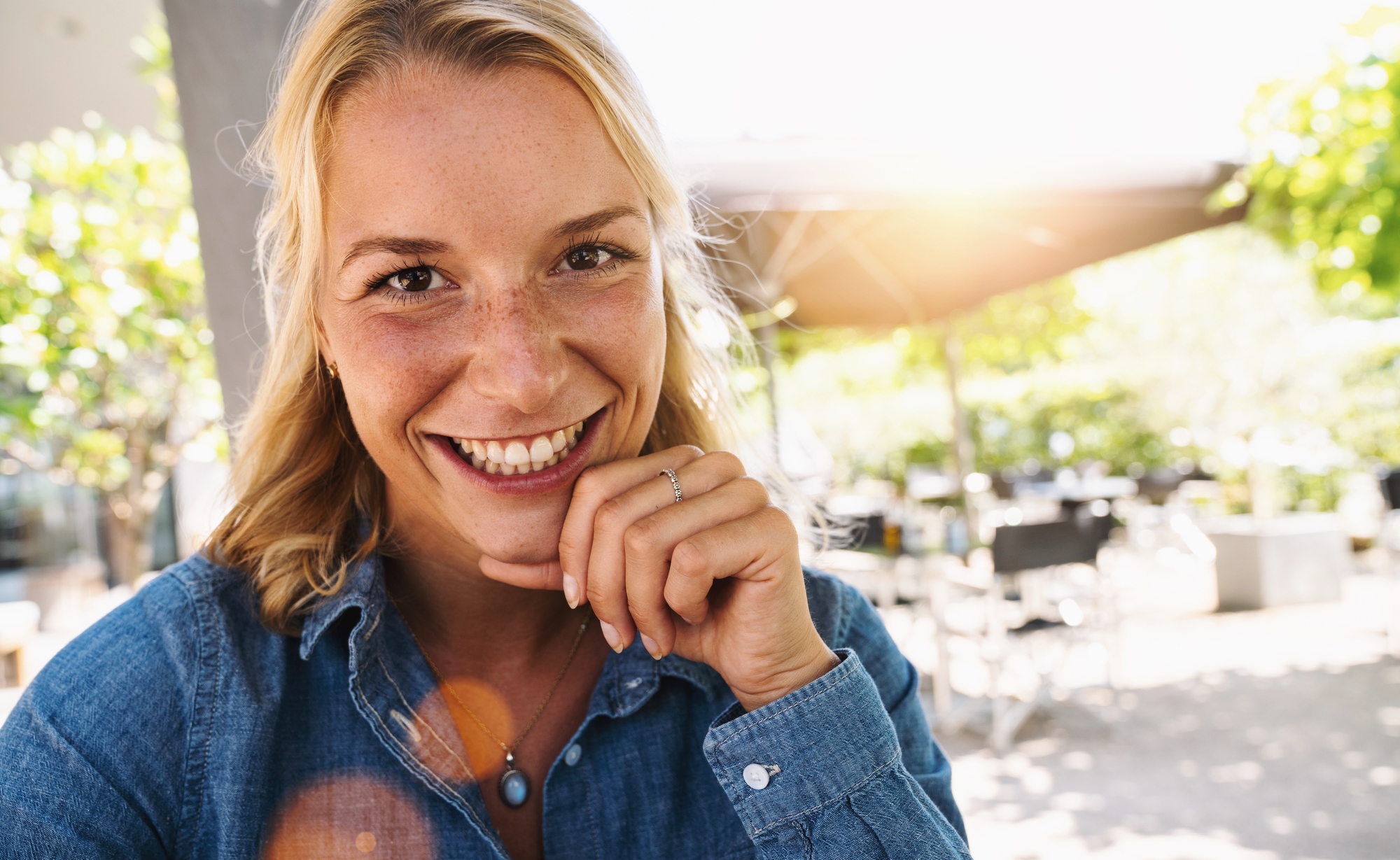 A woman with blonde hair smiles warmly at the camera. She's wearing a blue denim shirt and sitting outdoors, with sunlight filtering through green foliage in the background. She rests her chin on her hand, creating a relaxed, cheerful atmosphere.