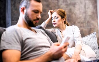 A woman leans on the back of a couch, looking at a man who is focused on his phone. They are sitting in a modern, comfortable living room. Both appear to be in casual clothing, and there is a sense of tension or concern in her expression.