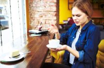 A woman sitting in a cafe next to a window looks puzzled while holding a cup of coffee. A slice of cheesecake is on the table in front of her. She appears confused or concerned as she examines the cup.