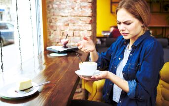 A woman sitting in a cafe next to a window looks puzzled while holding a cup of coffee. A slice of cheesecake is on the table in front of her. She appears confused or concerned as she examines the cup.