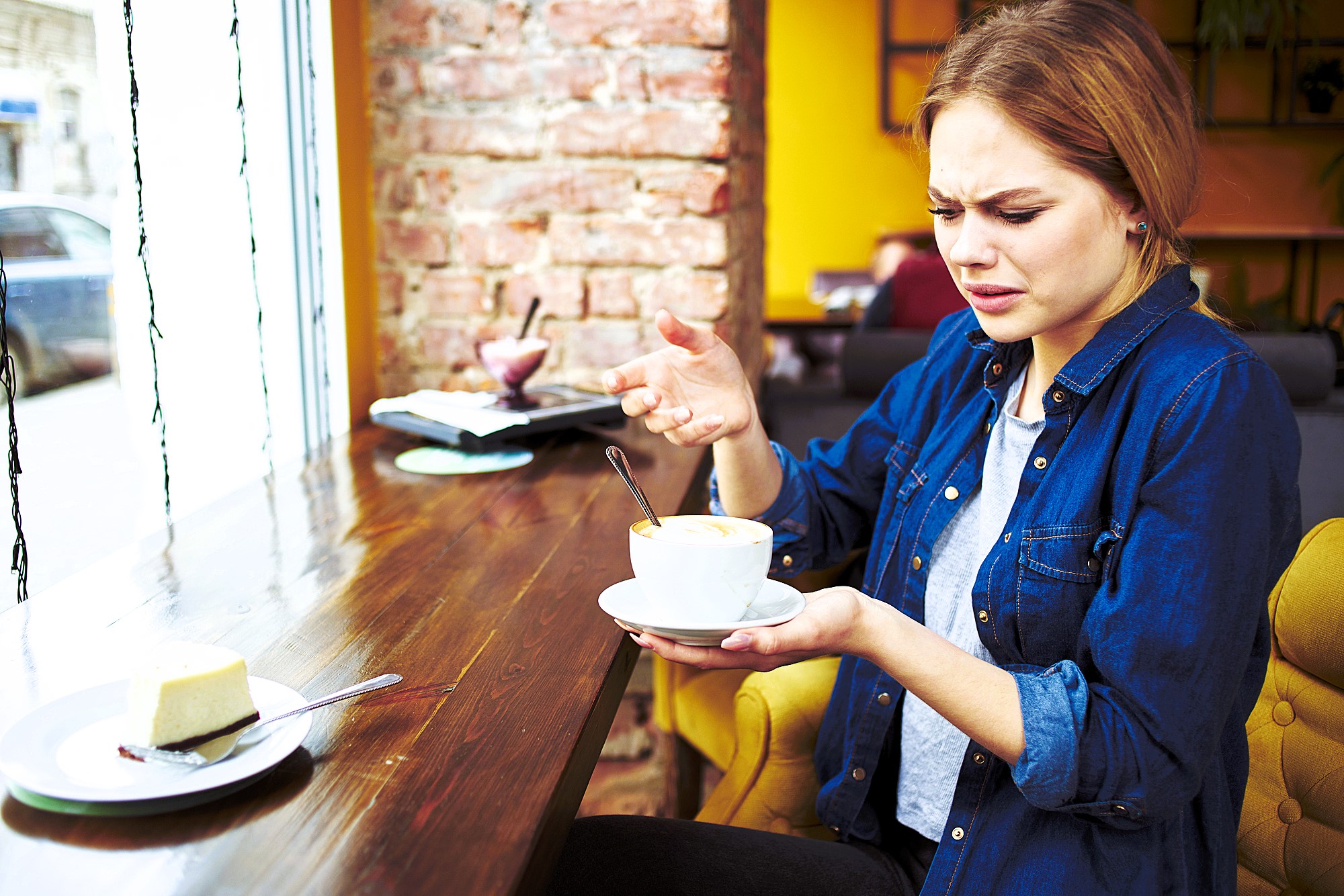 A woman sitting in a cafe next to a window looks puzzled while holding a cup of coffee. A slice of cheesecake is on the table in front of her. She appears confused or concerned as she examines the cup.