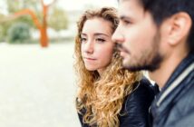 A woman with curly hair and a man with a beard sit closely together, both wearing black jackets. The woman gazes thoughtfully into the distance, while the man is slightly out of focus. They are outdoors with blurred greenery in the background.