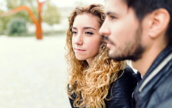 A woman with curly hair and a man with a beard sit closely together, both wearing black jackets. The woman gazes thoughtfully into the distance, while the man is slightly out of focus. They are outdoors with blurred greenery in the background.