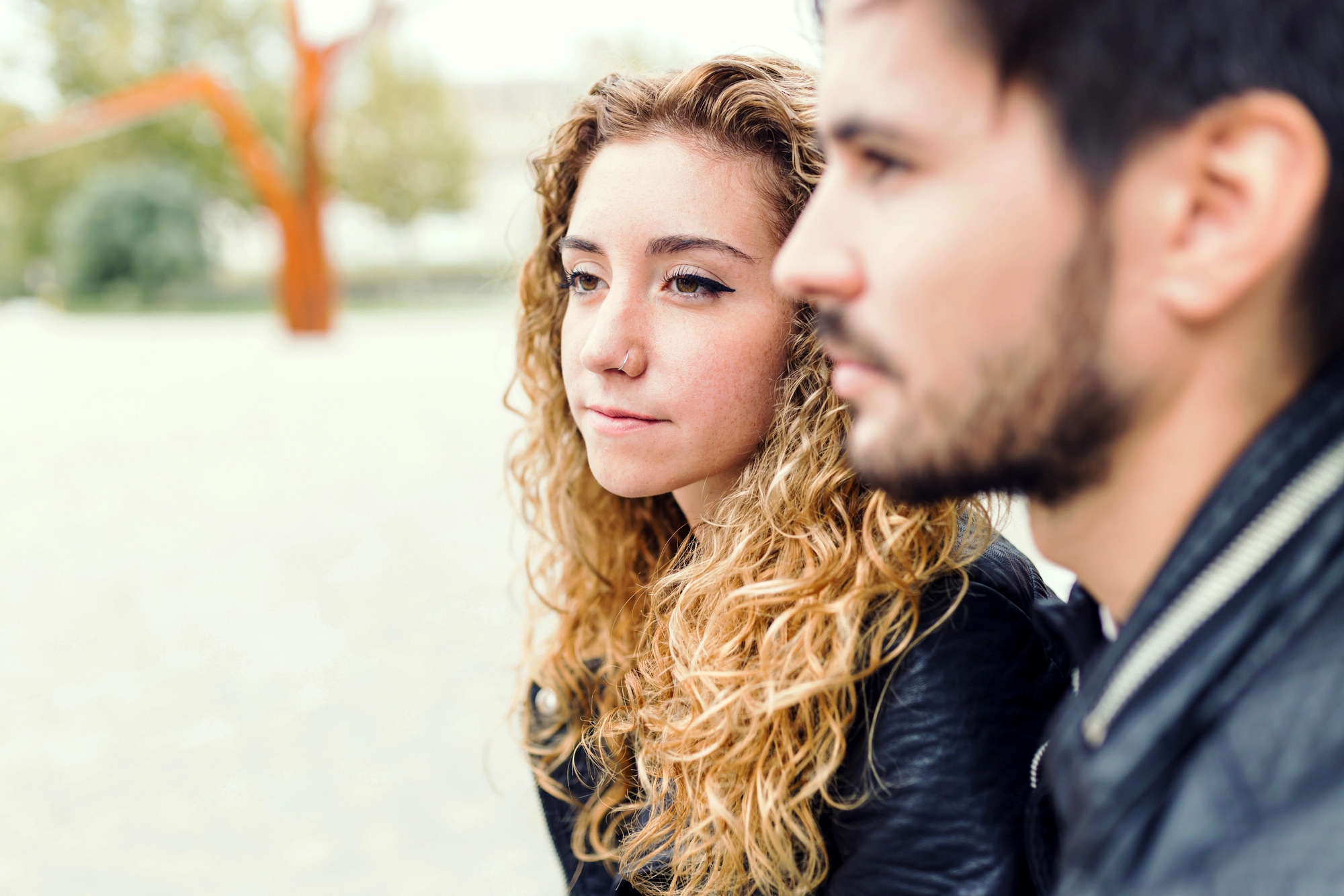 A woman with curly hair and a man with a beard sit closely together, both wearing black jackets. The woman gazes thoughtfully into the distance, while the man is slightly out of focus. They are outdoors with blurred greenery in the background.