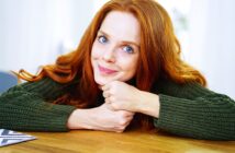 A person with long red hair and wearing a green sweater is resting their chin on their clasped hands on a wooden table, smiling at the camera. The background is softly blurred.