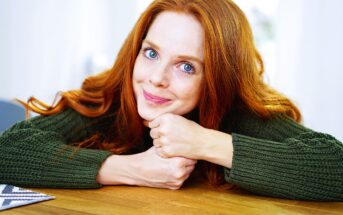 A person with long red hair and wearing a green sweater is resting their chin on their clasped hands on a wooden table, smiling at the camera. The background is softly blurred.