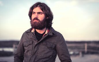 A bearded man with long hair stands outdoors, looking into the distance. He is wearing a dark jacket over a plaid shirt. The background is a soft-focus landscape with muted colors and a cloudy sky.