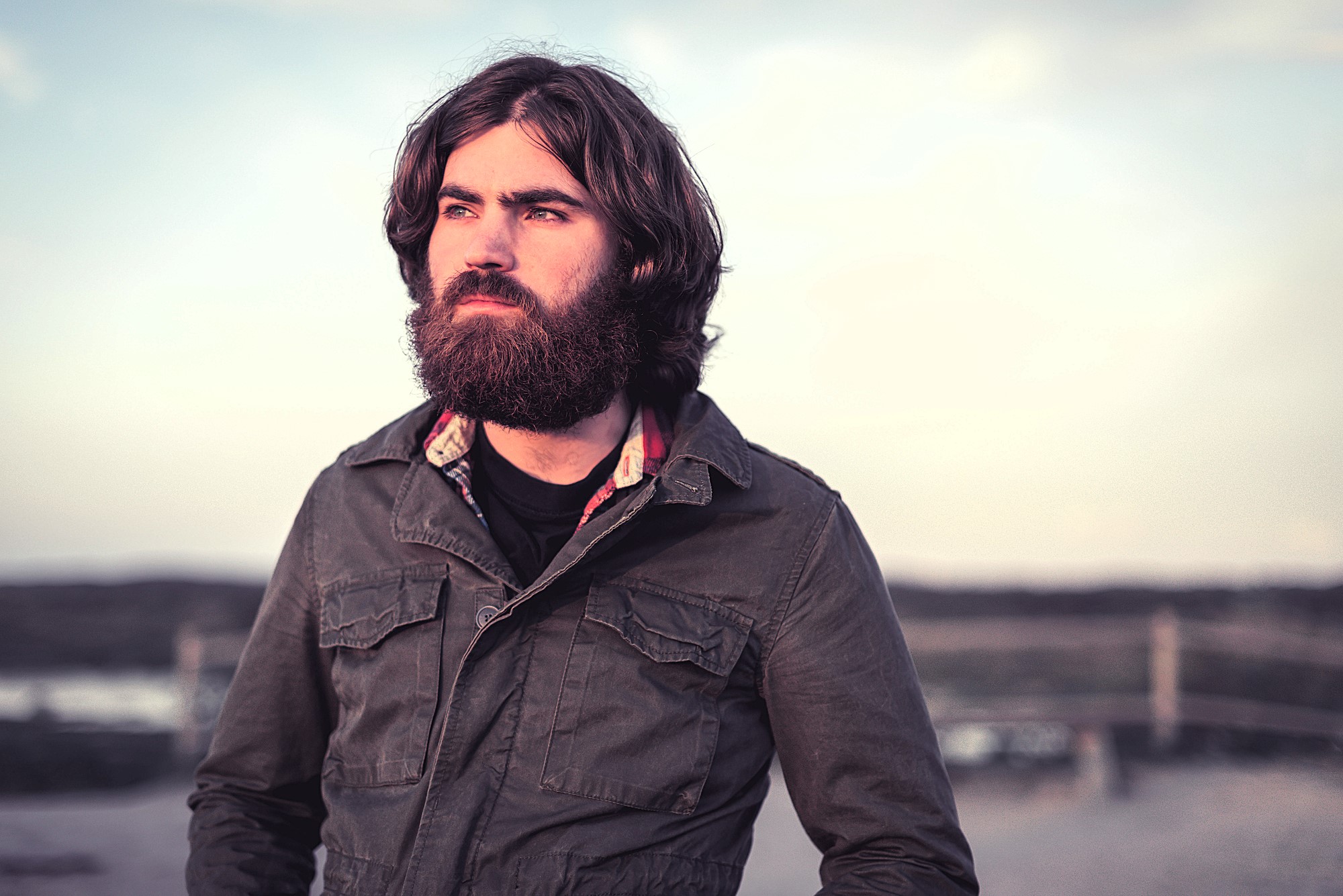 A bearded man with long hair stands outdoors, looking into the distance. He is wearing a dark jacket over a plaid shirt. The background is a soft-focus landscape with muted colors and a cloudy sky.