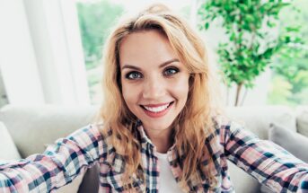 A woman with long blonde hair takes a selfie indoors. She is wearing a plaid shirt and smiling at the camera. Behind her, there's a large window with a view of greenery and a potted plant.