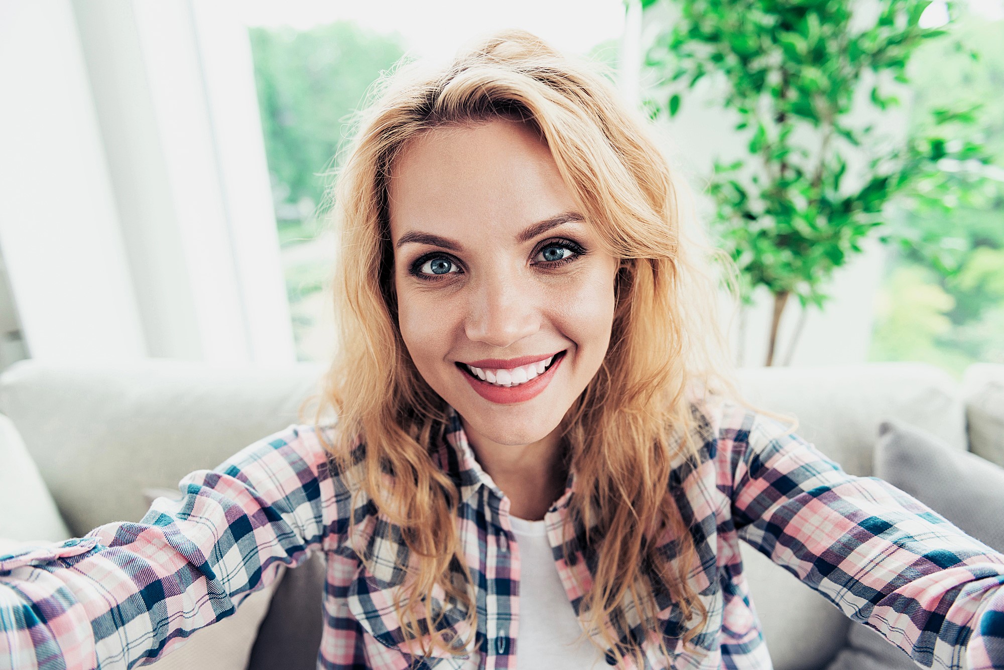 A woman with long blonde hair takes a selfie indoors. She is wearing a plaid shirt and smiling at the camera. Behind her, there's a large window with a view of greenery and a potted plant.