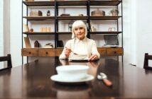 An elderly woman with white hair sits at the end of a long wooden dining table, holding a spoon in one hand. A bowl and spoon are in front of her. The background shows shelves with books and small decorative items.