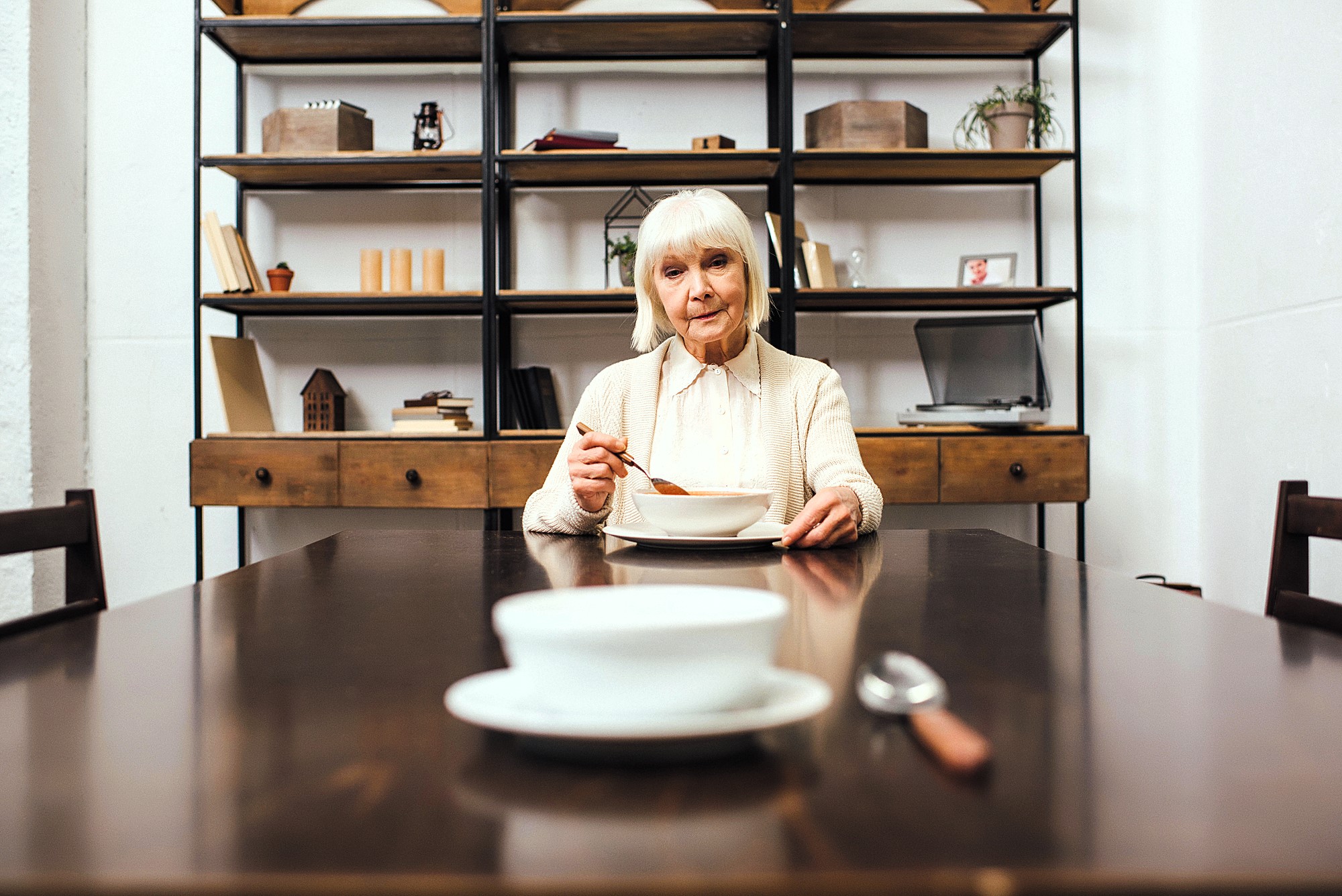 An elderly woman with white hair sits at the end of a long wooden dining table, holding a spoon in one hand. A bowl and spoon are in front of her. The background shows shelves with books and small decorative items.