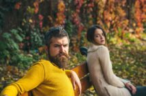 A man with a beard in a yellow sweater sits on a bench, looking serious, with autumn leaves in the background. A woman next to him in a cream sweater gazes at the camera. The scene is set in a park during fall.