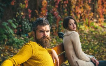 A man with a beard in a yellow sweater sits on a bench, looking serious, with autumn leaves in the background. A woman next to him in a cream sweater gazes at the camera. The scene is set in a park during fall.