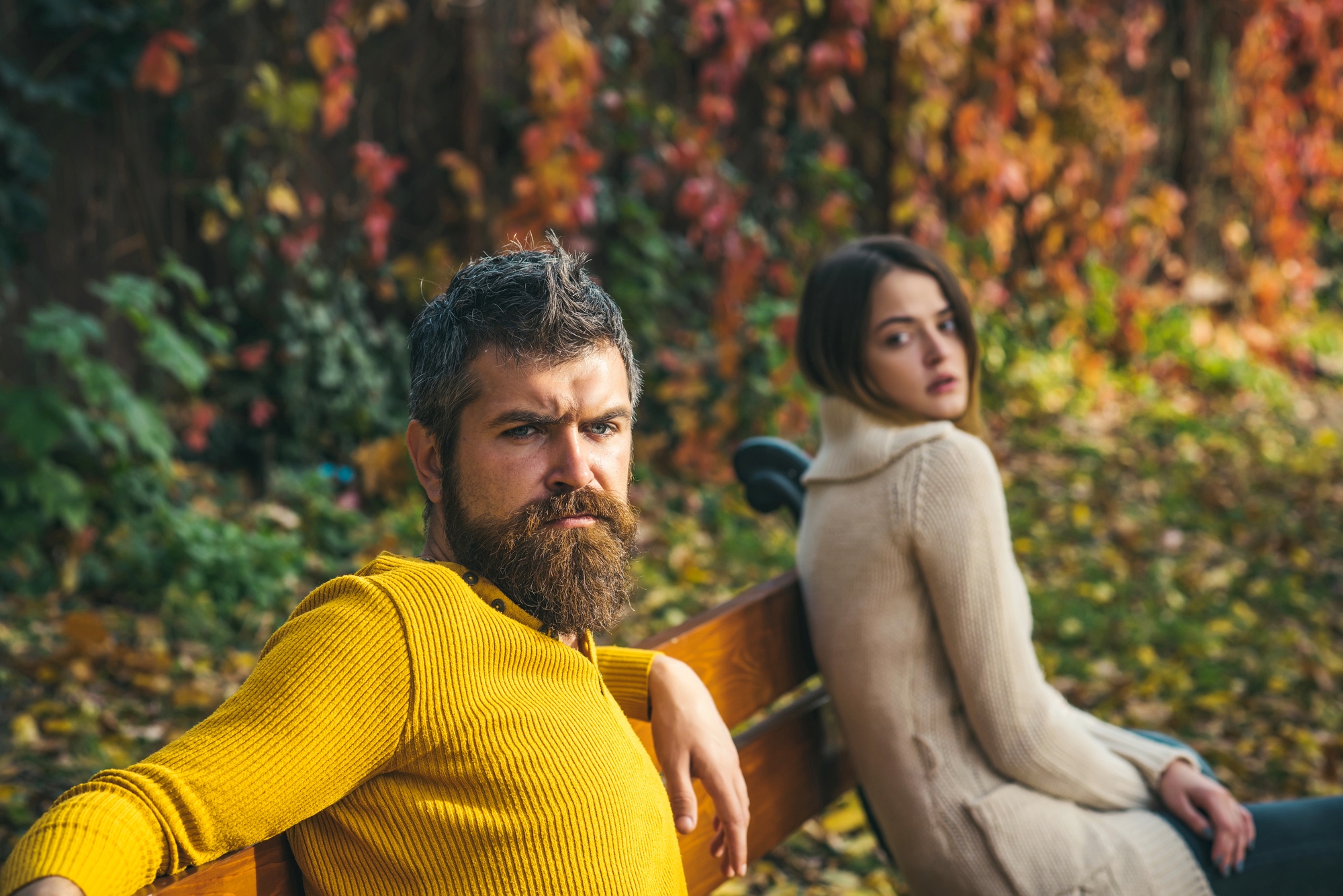 A man with a beard in a yellow sweater sits on a bench, looking serious, with autumn leaves in the background. A woman next to him in a cream sweater gazes at the camera. The scene is set in a park during fall.