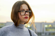 A person with shoulder-length brown hair and glasses stands outdoors, wearing a gray turtleneck. The background is a blurred cityscape with a soft, overcast sky. The individual looks slightly to the side with a neutral expression.