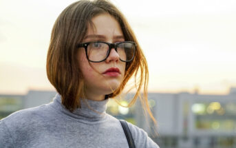 A person with shoulder-length brown hair and glasses stands outdoors, wearing a gray turtleneck. The background is a blurred cityscape with a soft, overcast sky. The individual looks slightly to the side with a neutral expression.