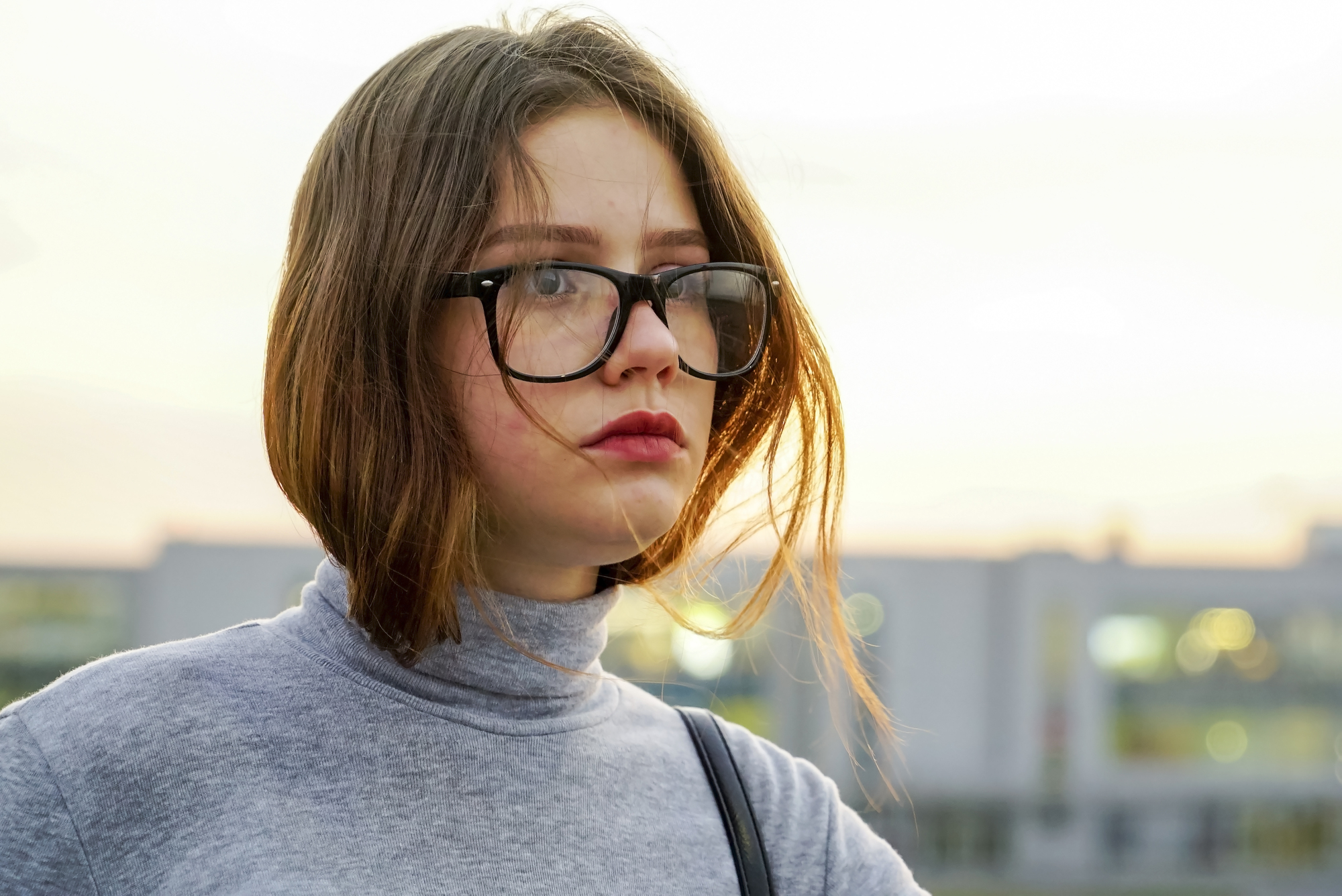 A person with shoulder-length brown hair and glasses stands outdoors, wearing a gray turtleneck. The background is a blurred cityscape with a soft, overcast sky. The individual looks slightly to the side with a neutral expression.