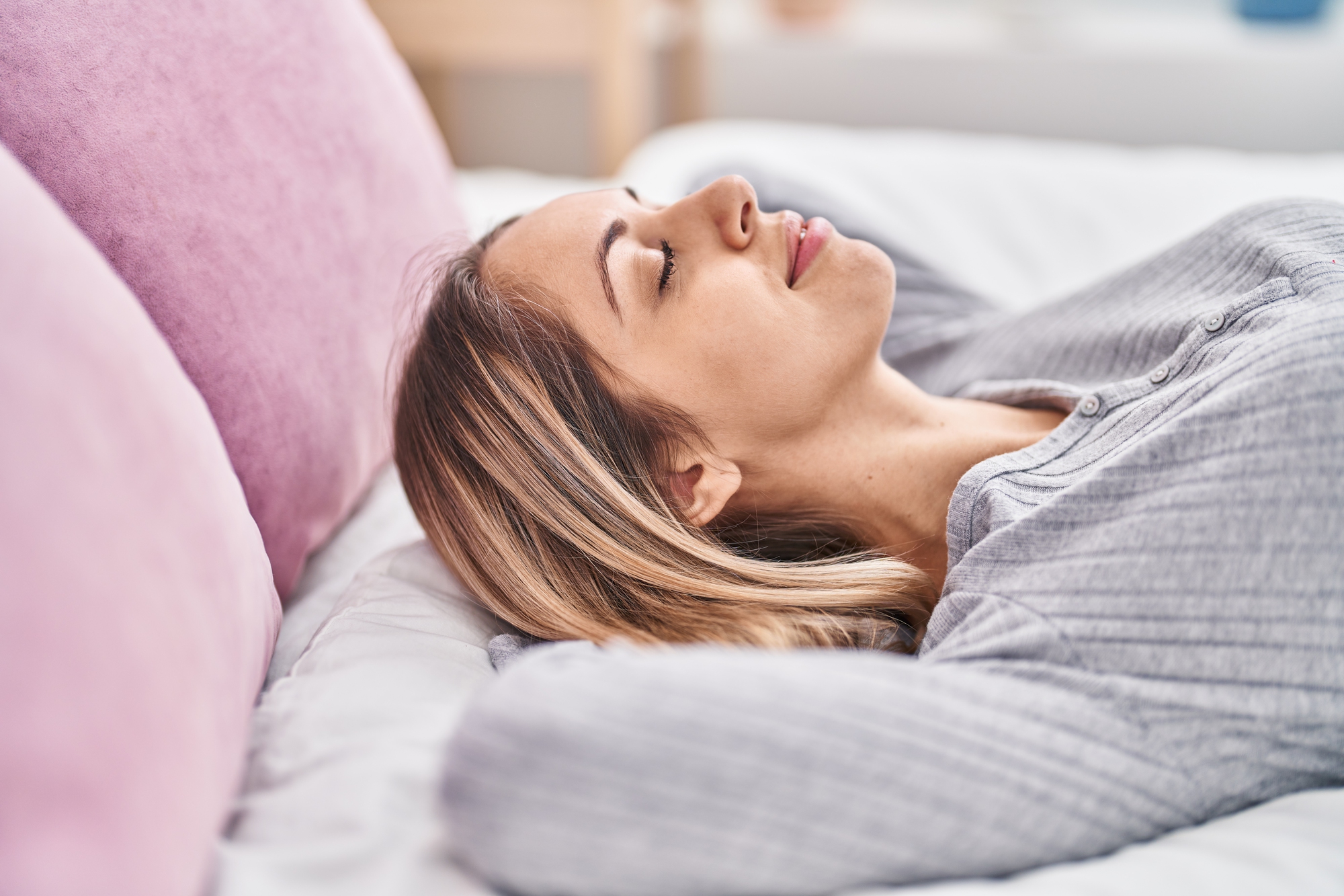 A woman with long hair relaxes on a bed with pink cushions, wearing a gray shirt. She is lying on her back with her hands behind her head, eyes closed, appearing calm and content.