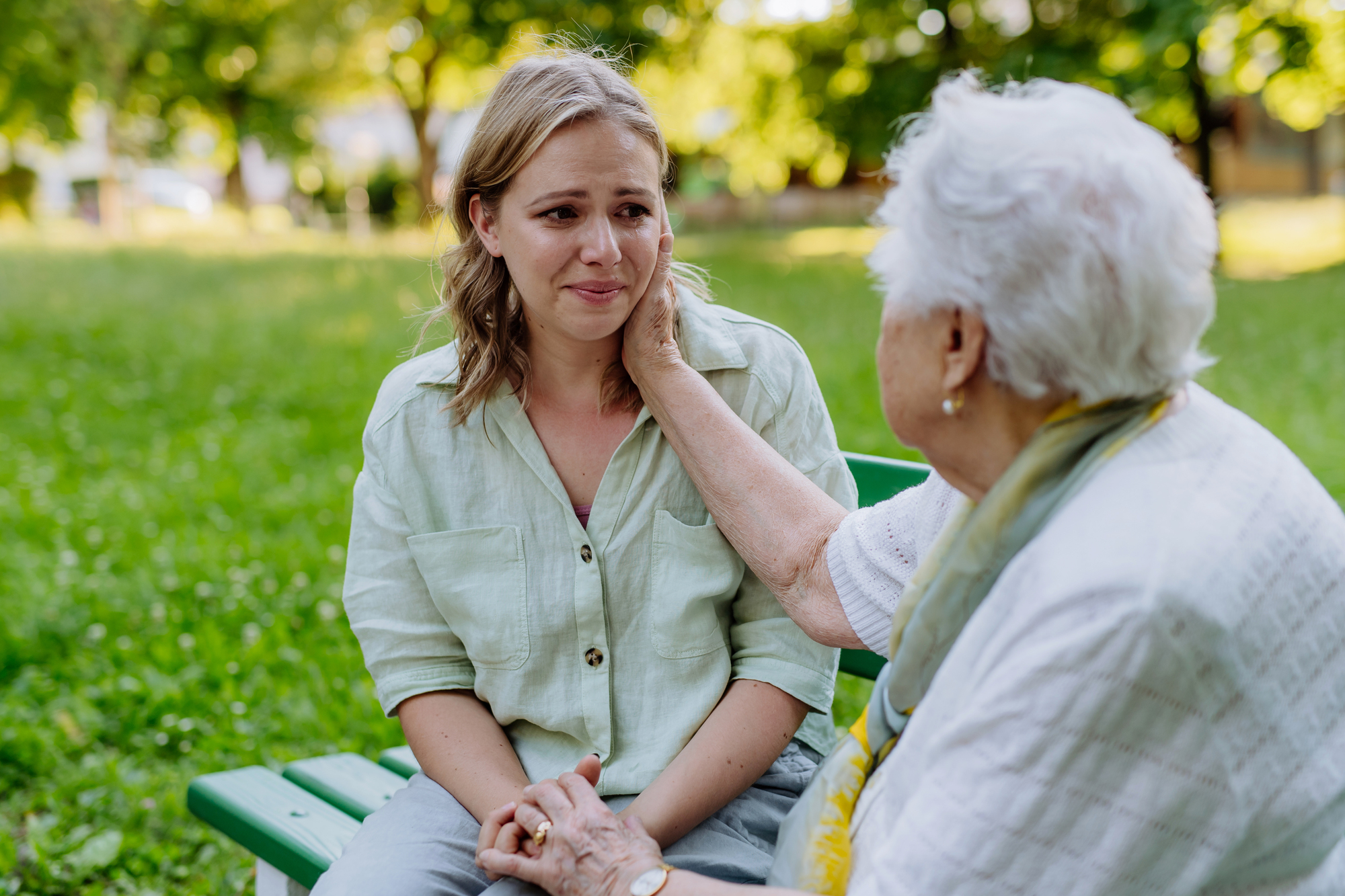 An older woman gently comforts a younger woman on a park bench. The younger woman appears emotional, while the older woman places a hand on her cheek. They are surrounded by green trees and grass, creating a serene outdoor setting.