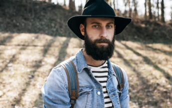 A bearded man wearing a black hat, denim jacket, and striped shirt stands outdoors. Sunlight filters through trees in the background, casting shadows on the ground.