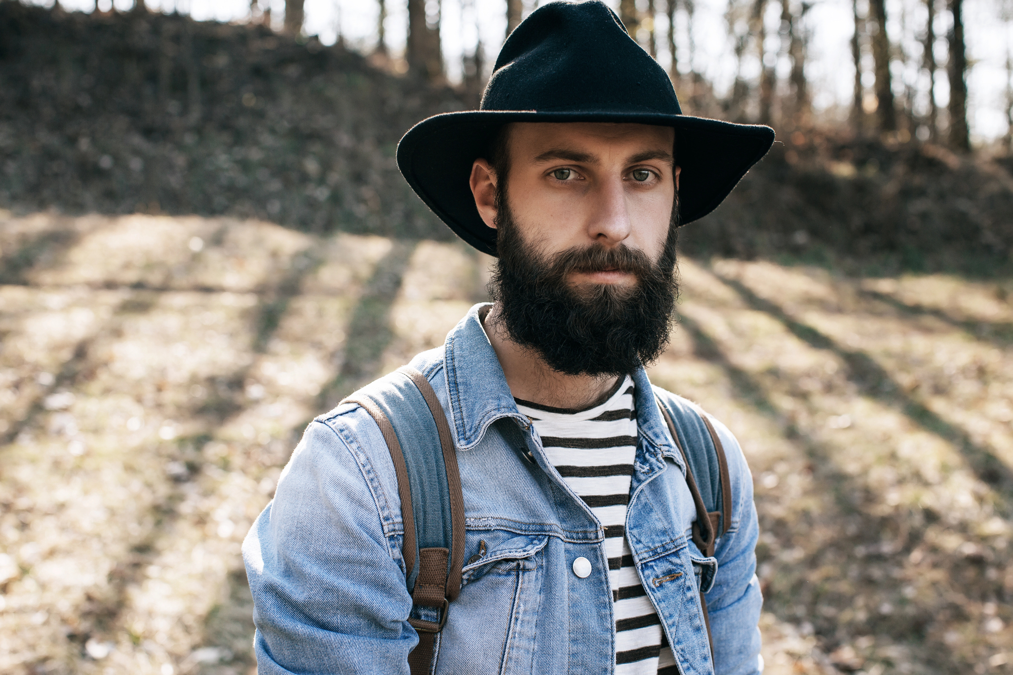 A bearded man wearing a black hat, denim jacket, and striped shirt stands outdoors. Sunlight filters through trees in the background, casting shadows on the ground.