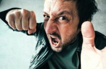 A man wearing a black leather jacket is expressing anger, with his mouth open and fist raised. His face is close to the camera, conveying a sense of frustration or confrontation. The background is a neutral gray.