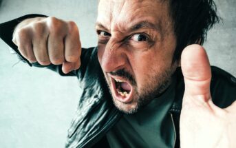 A man wearing a black leather jacket is expressing anger, with his mouth open and fist raised. His face is close to the camera, conveying a sense of frustration or confrontation. The background is a neutral gray.