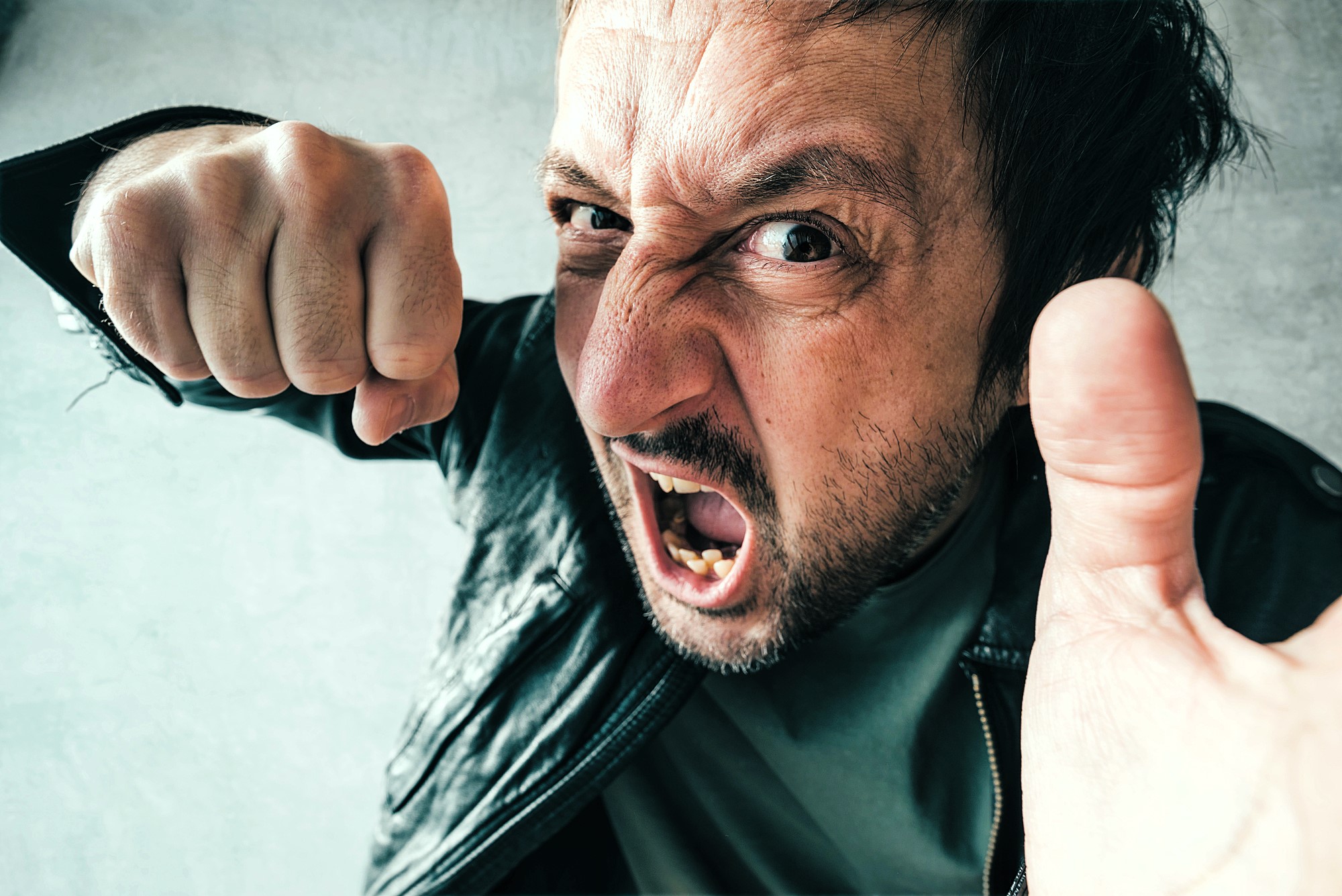 A man wearing a black leather jacket is expressing anger, with his mouth open and fist raised. His face is close to the camera, conveying a sense of frustration or confrontation. The background is a neutral gray.