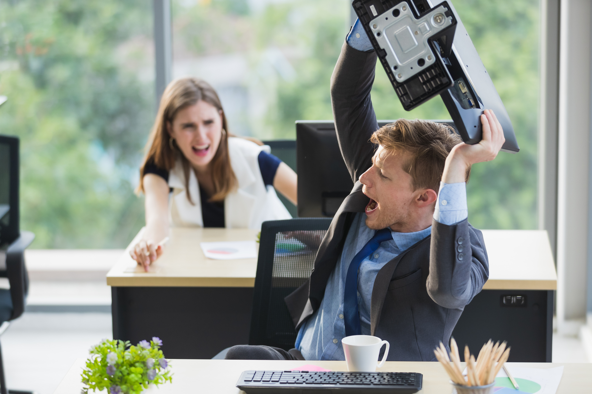 A man in a suit is angrily lifting a keyboard over his head, seated at a desk with a coffee cup and computer monitor. A woman in the background appears shocked, with her mouth open and hand raised. Large windows with greenery outside are visible.