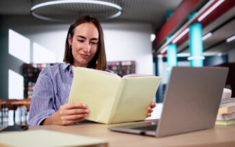 A person with shoulder-length hair is sitting at a library desk, reading a folder. They are in front of a laptop with books stacked nearby. The background includes shelves and a modern circular light fixture.