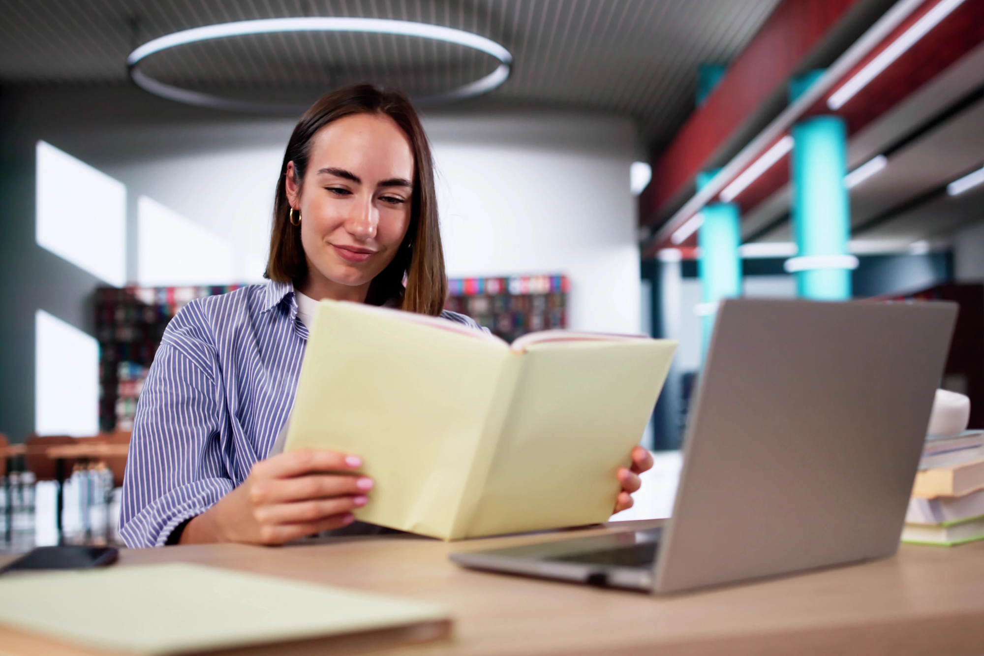 A person with shoulder-length hair is sitting at a library desk, reading a folder. They are in front of a laptop with books stacked nearby. The background includes shelves and a modern circular light fixture.