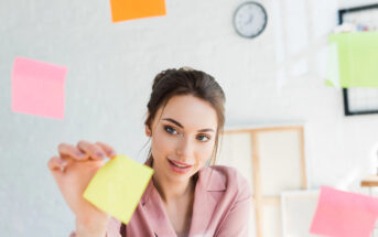 A woman in a pink shirt is smiling while interacting with colorful sticky notes floating around her in a bright, modern office. A wall clock and blurred background elements are visible in the room.