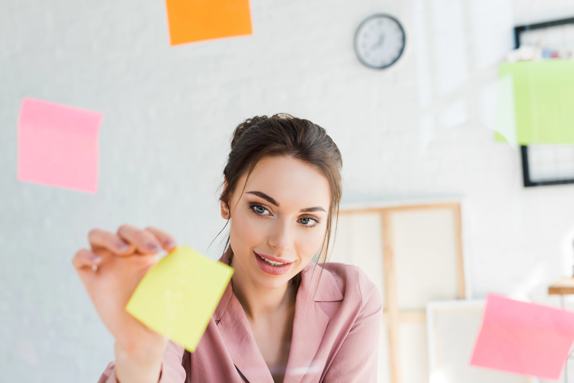 A woman in a pink shirt is smiling while interacting with colorful sticky notes floating around her in a bright, modern office. A wall clock and blurred background elements are visible in the room.