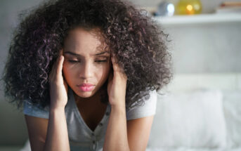 A woman with curly hair is sitting on a bed, holding her head with both hands. She appears pensive or stressed, wearing a casual gray shirt. The background includes a soft-focus white wall and shelf.