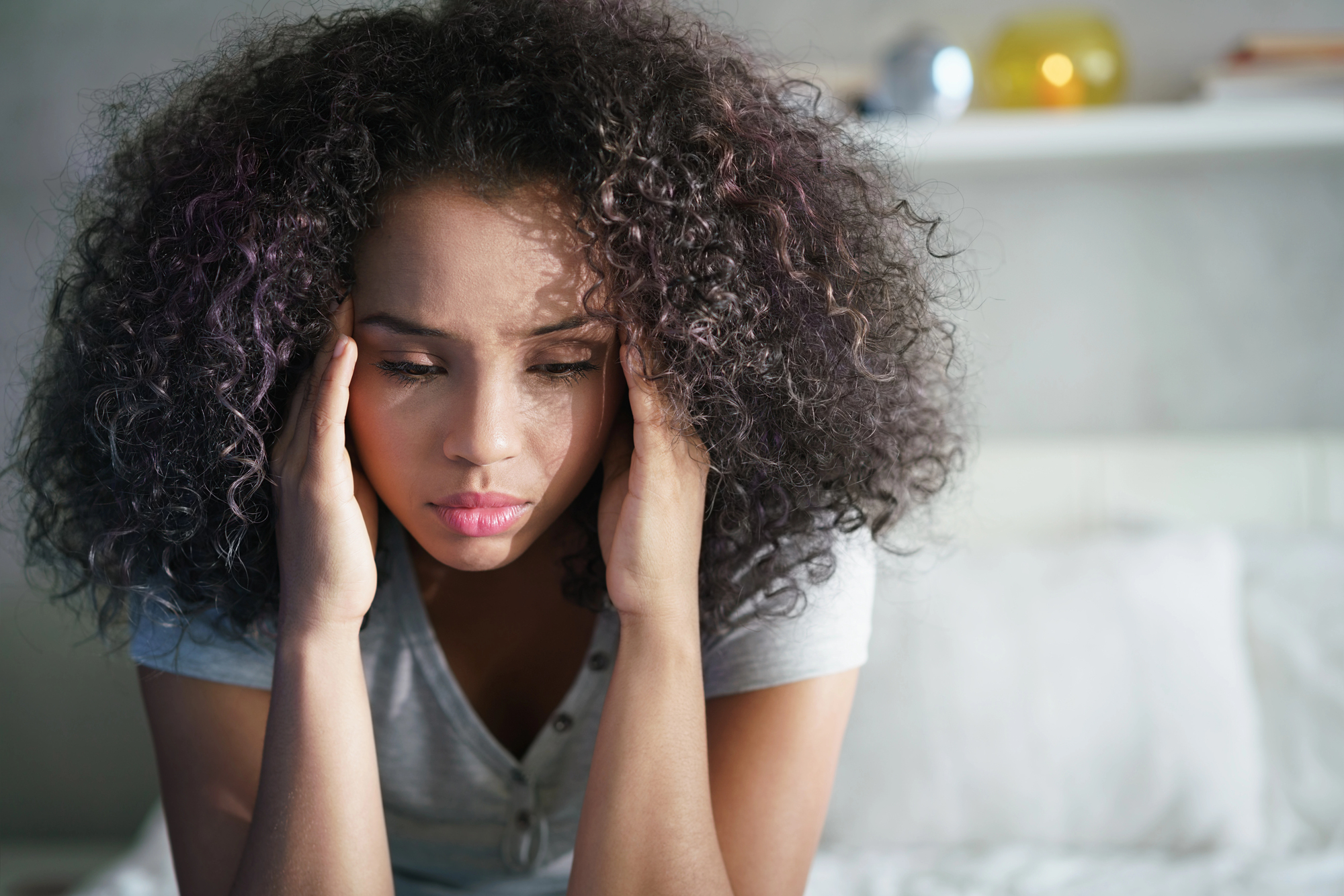 A woman with curly hair is sitting on a bed, holding her head with both hands. She appears pensive or stressed, wearing a casual gray shirt. The background includes a soft-focus white wall and shelf.