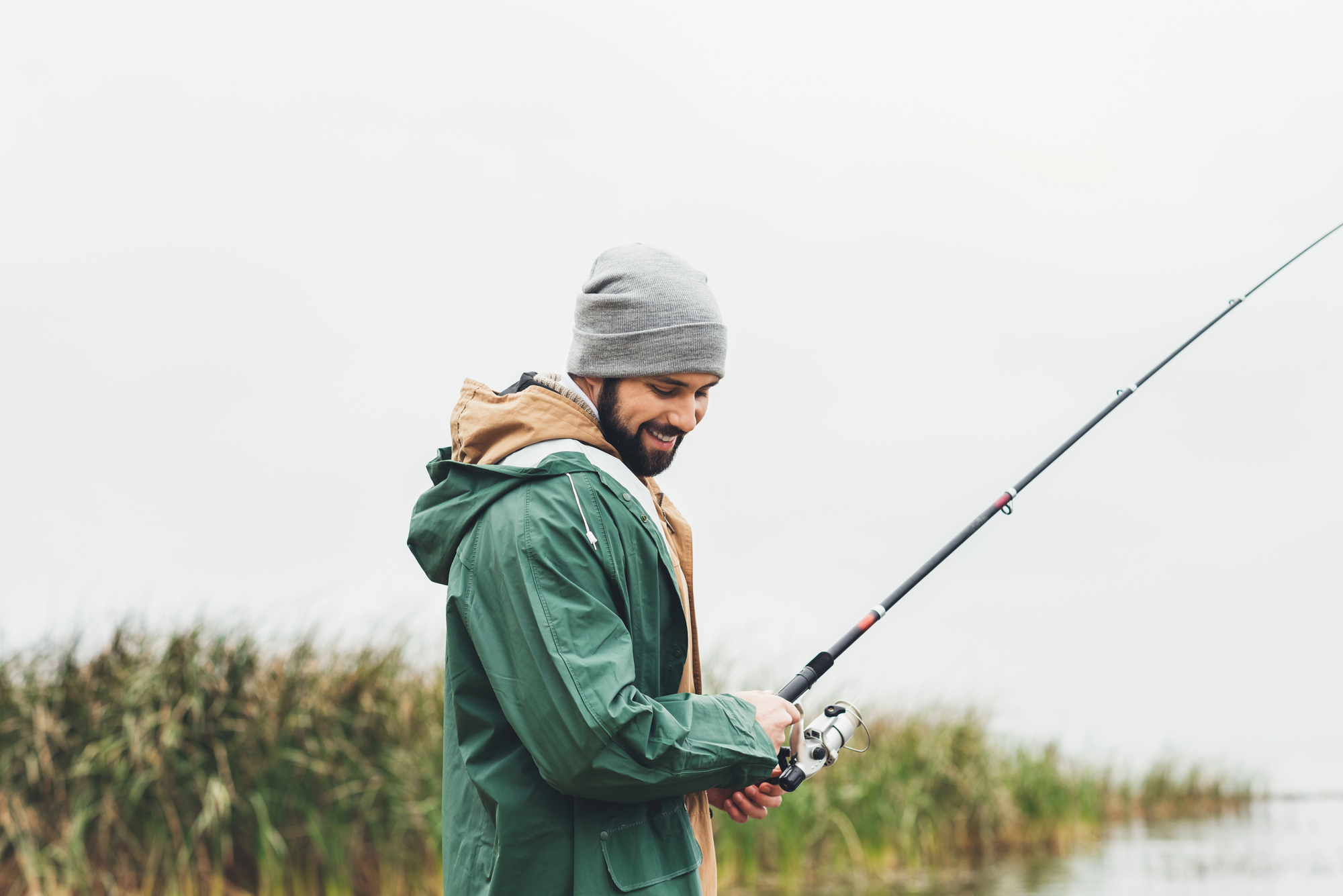 A person wearing a beanie and green jacket is holding a fishing rod, smiling at a lake or riverbank. There are reeds in the background and the sky is overcast.