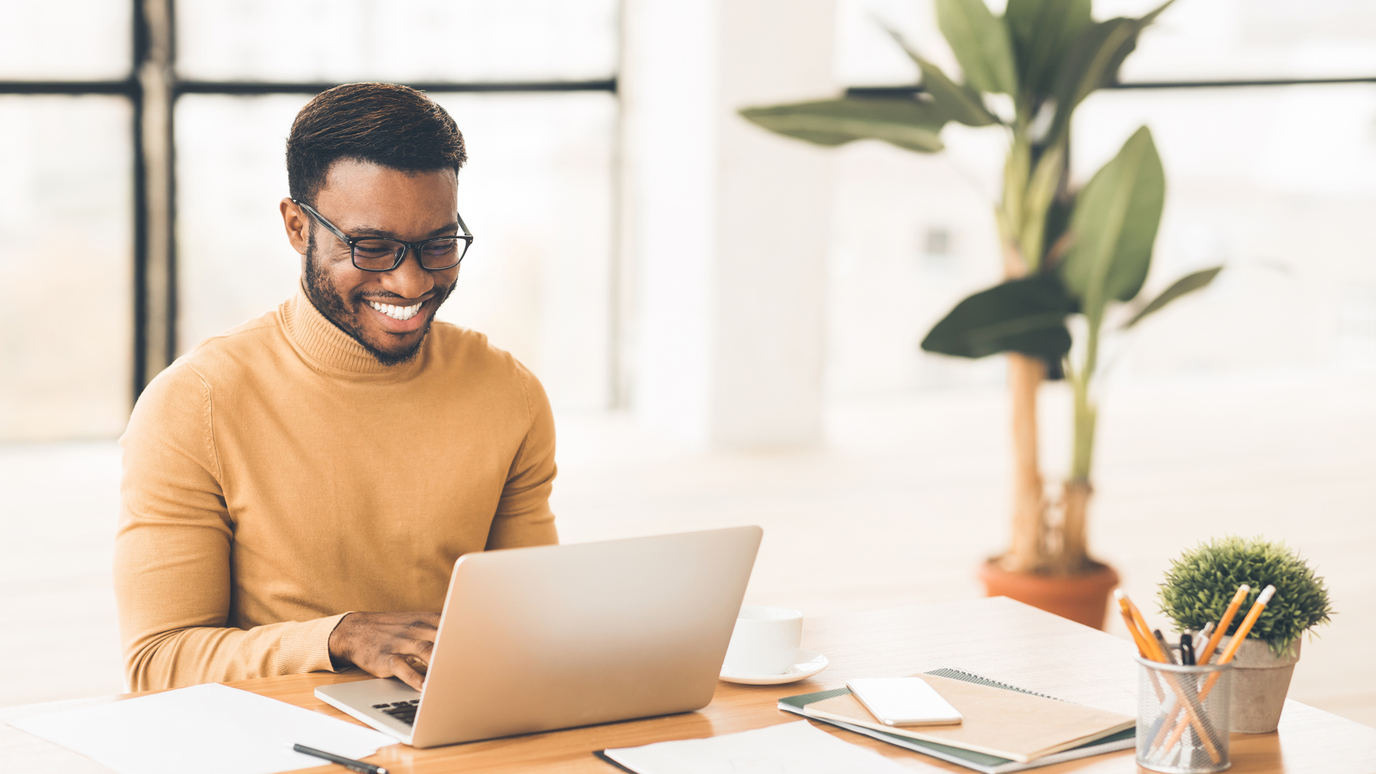 A person wearing glasses and a brown sweater is smiling while using a laptop at a desk. The desk has a small plant, a cup, and stationery. A large window and a tall plant are in the background, creating a bright and airy atmosphere.