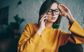 A person in a yellow sweater sits with their eyes closed, holding a phone to their ear and resting their other hand on their head, appearing thoughtful or tired. They wear glasses and have short, straight hair. The background is softly blurred.