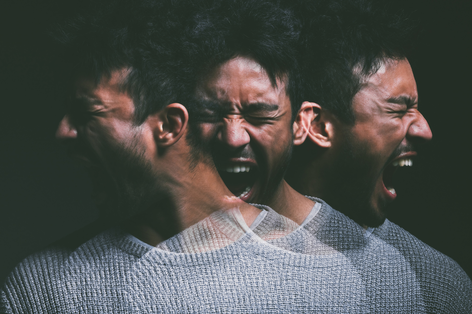 A single man with dark hair and a beard is captured in three overlapping, expressive positions. He is wearing a gray sweater and appears to be shouting or expressing strong emotions against a dark background.