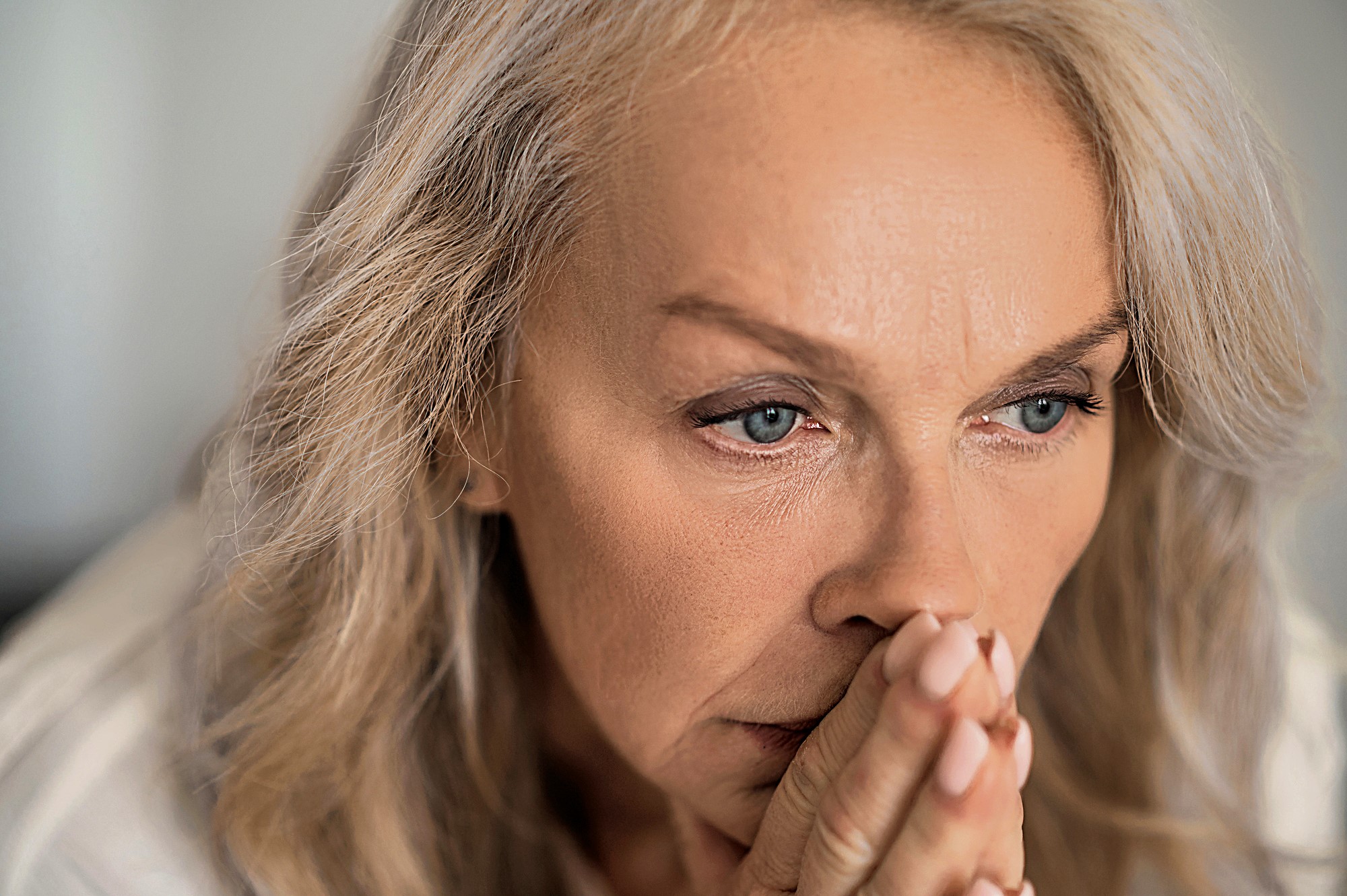 A close-up of a woman with light hair, looking contemplative. Her hands are clasped in front of her face, and she appears to be deep in thought. The background is soft and out of focus, emphasizing her expression.