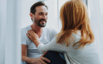 A man with a beard and a woman with red hair sit by a window, smiling and engaged in conversation. The man wears a white t-shirt and the woman has her hand on his shoulder. Natural light filters through sheer curtains.